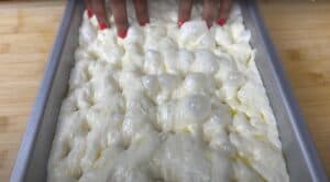 Fingers pressing into airy focaccia dough in a baking tray on a wooden surface.